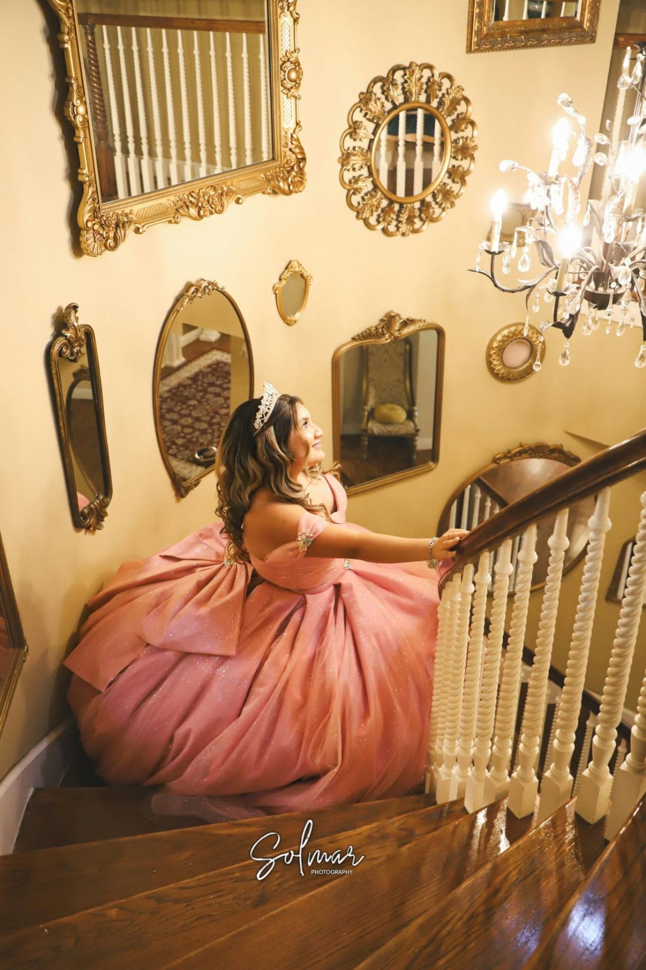 Quinceañera glancing up on the staircase beside crystal chandelier
